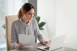 Woman typing on her laptop while referencing a sheet of paper